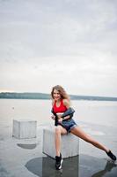 Sexy curly model girl in red top, jeans denim shorts, leather jacket and sneakers posed at stone cubes. photo