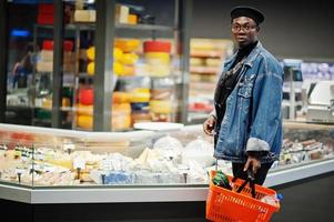 elegante hombre afroamericano casual con chaqueta de jeans y boina negra sosteniendo una cesta, parado cerca de la nevera de queso y comprando en el supermercado. foto