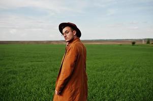 hombre elegante con gafas, chaqueta marrón y sombrero posado en campo verde. foto