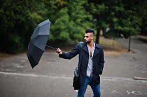 hombre de barba árabe alto de moda vestido con abrigo negro con paraguas y estuche posado en el día de la lluvia. foto