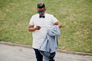 Stylish arabian man with jacket, sunglasses and bow tie posed outdoor and looking on mobile phone. Arab model boy. photo