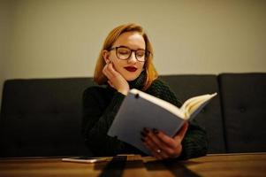 Cheerful young beautiful redhaired woman in glasses, wear on green warm wool sweater,  sitting at cafe and using her notebook. photo