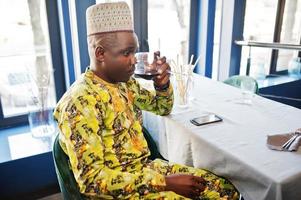 Friendly afro man in traditional yellow clothes and cap drinking red wine at restaurant. photo