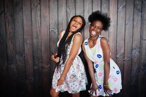 Two black african girlfriends at summer dresses posed against dark wooden background and showing tongue. photo