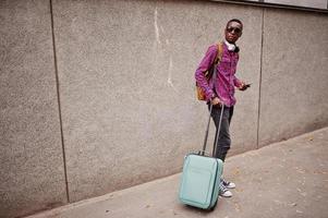 African american man in checkered shirt, sunglasses and earphones with suitcase. Black man traveler against wall holding mobile phone. photo