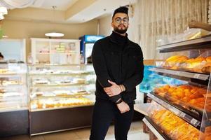 Arab man wear on black jeans jacket and eyeglasses in cafe choose bakery. Stylish and fashionable arabian model guy. photo