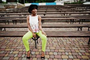 Amazing african american model woman in green pants and black hat posed at bench. photo