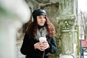 Curly mexican girl in leather cap and plastic cup of coffee at hand walking at streets of city. photo