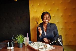 African american businesswoman eating cheese pizza in cafe. Black girl having rest. photo