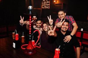 Group of indian friends having fun and rest at night club, drinking cocktails and smoke hookah. Hands with two fingers up. photo