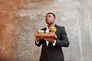 Respectable young african american man in black suit hold tray with double burger against gray wall. He breathes smell of a delicious hamburger.