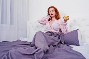 Gorgeous red haired girl in pink blouse and red skirt with glass of wine at hand on the bed at room. photo