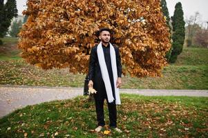 Indian stylish man in black traditional clothes with white scarf posed outdoor against yellow autumn. photo