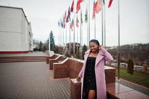 joven y elegante mujer afroamericana en la calle, con abrigo de moda y anteojos, contra banderas de diferentes países del mundo. foto