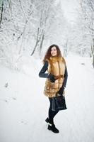 Elegance curly girl in fur coat and handbag at snowy forest park at winter. photo