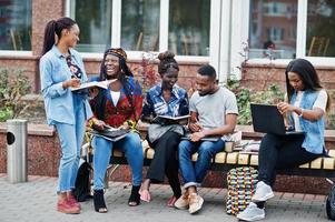 Group of five african college students spending time together on campus at university yard. Black afro friends studying at bench with school items, laptops notebooks. photo