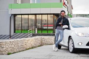 South asian man or indian male wear red eyeglasses stand near his white transportation on car wash. photo