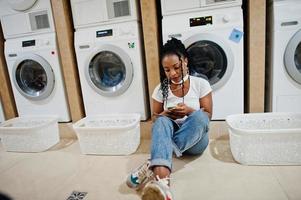 alegre mujer afroamericana sentada con auriculares y leyendo una revista cerca de la lavadora en la lavandería de autoservicio. foto