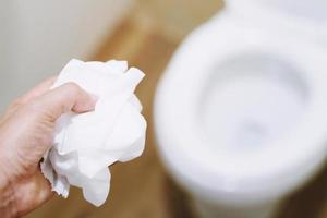 Primer plano de un joven tirando una toallita húmeda al inodoro, en un baño de azulejos blancos foto