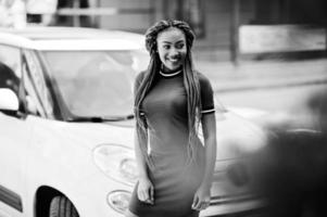 linda y delgada chica afroamericana vestida de rojo con rastas posadas al aire libre en el fondo de la calle pequeño coche blanco. elegante modelo negro. foto