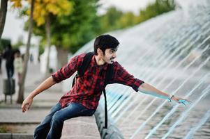 Young indian student man at checkered shirt and jeans with backpack posed on evening city against fountains. photo