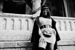African american woman at violet dress and cap posed outdoor against old column with red curtains. photo