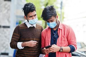 Coronavirus covid-19 concept. Two south asian indian man wearing mask for protect from corona virus looking at mobile phone. New normal lifestyle post pandemic in India.