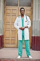 Stylish african american doctor with stethoscope and lab coat posed against door of hospital. photo