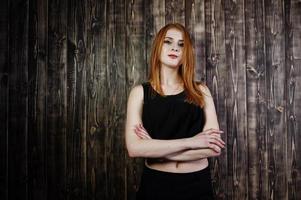 Portrait of a beautiful redheaded girl in black top and black skirt posing in the studio next to the wooden wall. photo