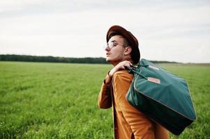 Stylish man in glasses, brown jacket and hat with bag posed on green field. photo