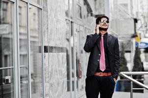 Young indian man on suit, tie and sunglasses posed outdoor, speaking on phone. photo