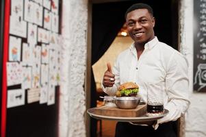 Young african american waiter man hold tray with burger at restaurant and show thumb up. photo