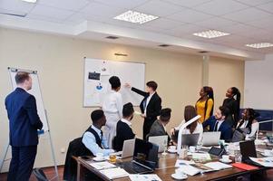 Mixed race business coach presenting report standing near whiteboard pointing on sales statistic shown on diagram and chart teach diverse company members gathered together in conference room. photo