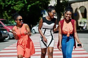 Three stylish african american womans walking on crosswalk or pedestrian crossing, speaking each other and having fun. photo