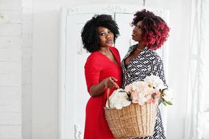 dos mujeres afroamericanas de moda vestidas de noche de pie con una cesta de flores en las manos contra el viejo armario vintage en la habitación blanca. foto