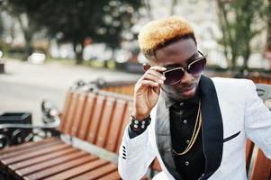 Chic handsome african american man in white suit and sunglasses sitting on bench. photo