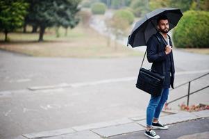 hombre de barba árabe alto de moda vestido con abrigo negro con paraguas y estuche posado en el día de la lluvia. foto