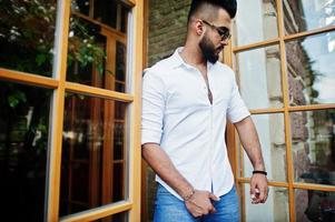 Stylish tall arabian man model in white shirt, jeans and sunglasses posed at street of city. Beard attractive arab guy against entrance door of restaurant. photo