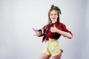 joven ama de casa divertida en camisa a cuadros y pantalones cortos amarillos pin up estilo con cacerola y cuchara de cocina aislado sobre fondo blanco. foto