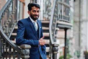 hombre indio de barba elegante con bindi en la frente, vestido con traje azul posado al aire libre contra escaleras de hierro y mostrando el pulgar hacia arriba. foto