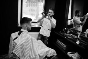 Young bearded man getting haircut by hairdresser while sitting in chair at barbershop. Barber soul. Master making photo on phone.