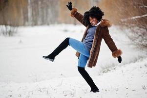 Mujer afroamericana de pelo rizado con abrigo de piel de oveja y guantes posados en el día de invierno, divirtiéndose y saltando. foto