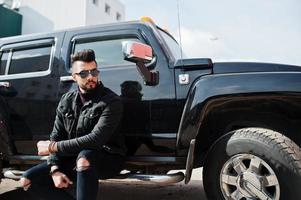 Fashion rich beard Arab man wear on black jeans jacket and sunglasses posed against big black suv car. Stylish, succesful and fashionable arabian model guy. photo
