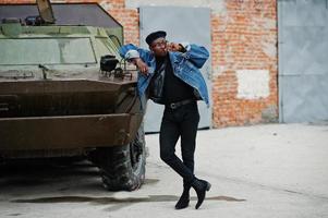 African american man in jeans jacket, beret and eyeglasses, with cigar posed against btr military armored vehicle. photo