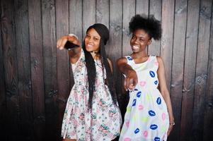 dos novias africanas negras con vestidos de verano posaron contra un fondo de madera oscura y mostraron los dedos a la cámara. foto