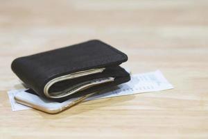 dinero y tarjeta de crédito en una cartera de cuero sobre una mesa de madera con teléfono inteligente móvil y fondo de papel de billete. Compra en línea. foto