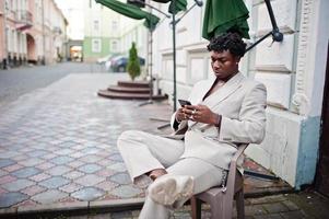 Stylish afro man in beige old school suit sitting outdoor in chair with mobile phone at hands. Fashionable young African male in casual jacket on bare torso. photo