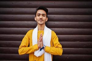 hombre indio elegante vestido con ropa tradicional amarilla con bufanda blanca posada al aire libre contra un fondo de madera y muestra el signo de las manos de namaste. foto