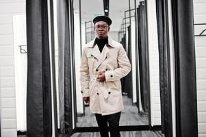 Stylish casual african american man at black beret and white trench raincoat at fitting room clothes store. photo