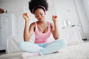 Young african american woman in pink singlet listen music on earphones at her white room. photo